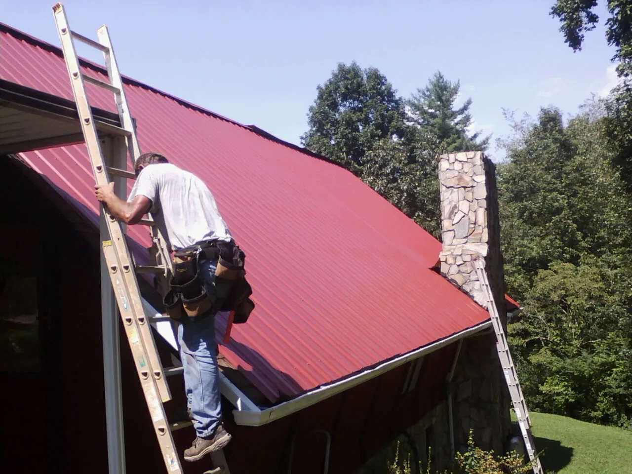 Roofer on a ladder beside a red metal roof and stone chimney — Soffit & Fascia Repair by Roofing Brothers in Cedar Rapids
